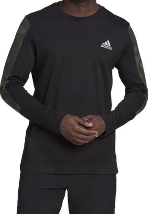 Hombre vistiendo una camiseta de manga larga negra de Adidas con rayas verde oliva oscuro en las mangas y el logo de Adidas en el pecho izquierdo, con las manos entrelazadas al frente.
