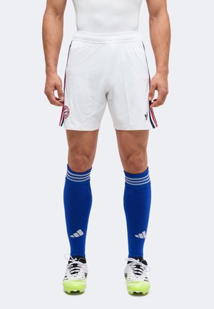 Homme portant un short de sport blanc avec des bandes latérales et le logo du club, des chaussettes hautes bleues et des chaussures de football blanches avec des semelles jaune néon.