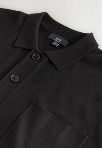 Polo noir en maille avec col, deux boutons noirs, poche avant et étiquette de marque visible à l'intérieur de l'encolure.