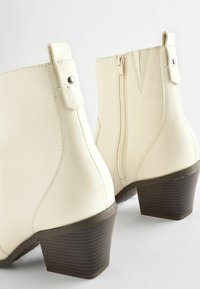Bottes de cheville crème avec une finition synthétique lisse, dotées d'une fermeture éclair latérale, de brides décoratives et d'un talon empilé marron.