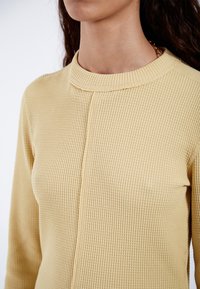 Light yellow ribbed knit top with a rounded neckline and long sleeves; features a centered seam detail on the front.