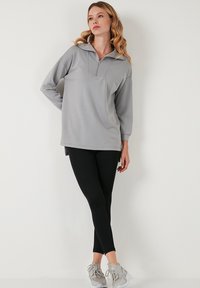 Siva zip-up jopica z dolgimi rokavi, ki ima visok ovratnik in stranske razpoke, v kombinaciji z črnimi legginsy in sivimi športnimi copati.
