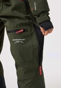 Grön vattentät jacka med zipficka med röda detaljer, meshfoder och varumärkeslogga. Ärmsluten har elastiska stängningar för komfort.