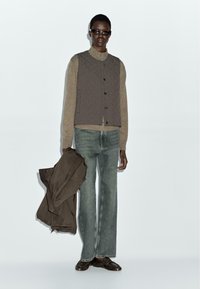 Gilet marrone trapuntato sopra una maglietta beige, abbinato a jeans in denim chiaro a gamba larga. Il modello tiene una giacca marrone; indossa occhiali da sole scuri.