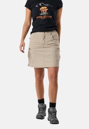 Frau trägt ein schwarzes Outdoor-T-Shirt, einen beigen Wanderrrock mit Taschen, schwarze Socken und graue Wanderschuhe vor einem einfarbigen Hintergrund.