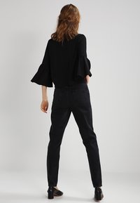 Blusa nera con maniche a campana, abbinata a jeans neri a vita alta. Indossata con scarpe nere con tacco. Modella vista da dietro.