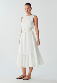 White sleeveless midi dress features a gathered waist and tiered ruffle hem. Smooth fabric with a flowing silhouette. Worn with black sandals.