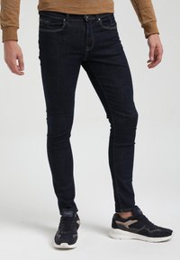 Hombre con jeans ajustados de color azul oscuro y zapatillas negras con suelas blancas, de pie contra un fondo claro y liso.
