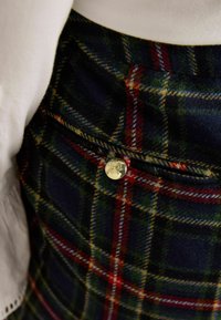 Pledēts svārks tumši zaļā un tumši zilā krāsā ar sarkanām un dzeltens krāsas akcentiem. Piedāvā zelta pogu detaļu un teksturētu vilnas maisījuma audumu.