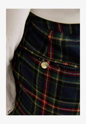 Pledinė sijonas tamsiai žalias ir tamsiai mėlynas su raudonomis ir geltonomis detalėmis. Yra auksinio mygtuko detalė ir tekstūruotas vilnos mišinio audinys.