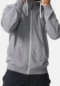 Personne portant un sweat à capuche gris à fermeture éclair avec des cordons blancs et un pantalon noir, ajustant la capuche près du cou.