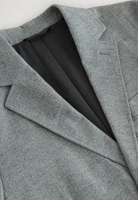 Sivi herringbone blazer s strukturalnim ovratnikom, koji pokazuje glatku crnu podstavu ispod. Značajna tekstura i krojačke detalje.