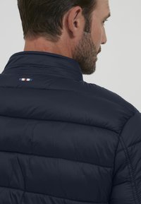 Marineblå puffjakke med høy krage og horisontal quiltedesign. Har en liten tri-farge aksent på bakhalsområdet.