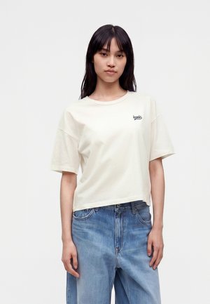 Tânără cu păr drept și închis la culoare, purtând un tricou alb oversized cu text "iconic" și blugi albaștri largi, pe un fundal simplu.