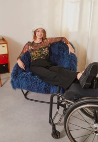 Blå, fluffy, overdimensjonert stol med metalldesign, ledsaget av en svart rullestol. Modellen har på seg en fargerik mønstrete topp og svarte bukser.