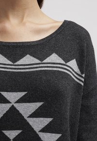 Pull gris avec un motif géométrique en triangle gris clair, doté d'un décolleté large en forme de scoop et d'une texture textile douce.