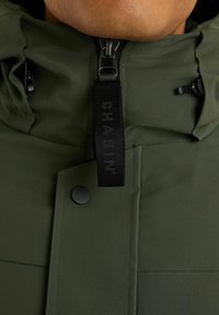 Giacca verde resistente all'acqua con collo alto, cerniera frontale e chiusura a scatto. Presenta un'etichetta in tessuto nero con logo e un piccolo dettaglio a tasca.