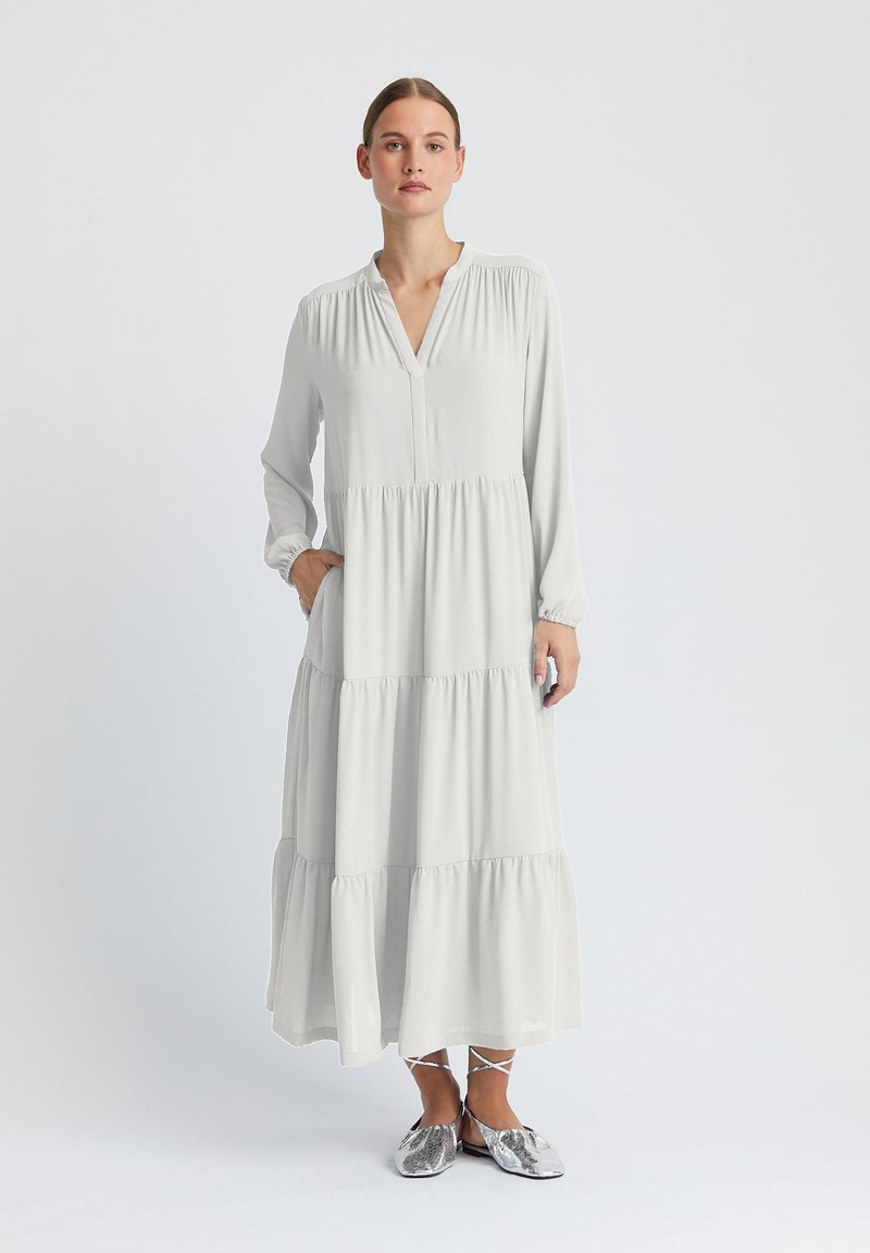 Robe longue gris clair en tissu lisse avec un col en V, des épaules froncées, un design à étages et des poignets élastiques. Le modèle porte des chaussures argentées.