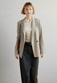 Blazer xadrez em bege e castanho, combinado com uma camisa polo bege clara. Saia de ganga escura, saco castanho e bijuteria minimalista em prata.