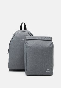 Šedý látkový batoh s zaobleným designem, který má přední zipovou kapsu a nastavitelné popruhy; vedle něj je odpovídající obdélníková taška s klopou.