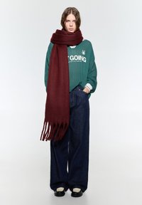 Vinskapad scarf i mjukt tyg med fransade ändar, kombinerad med en grön ribbad tröja och lösa mörkblå jeans. Svarta skor kompletterar outfiten.