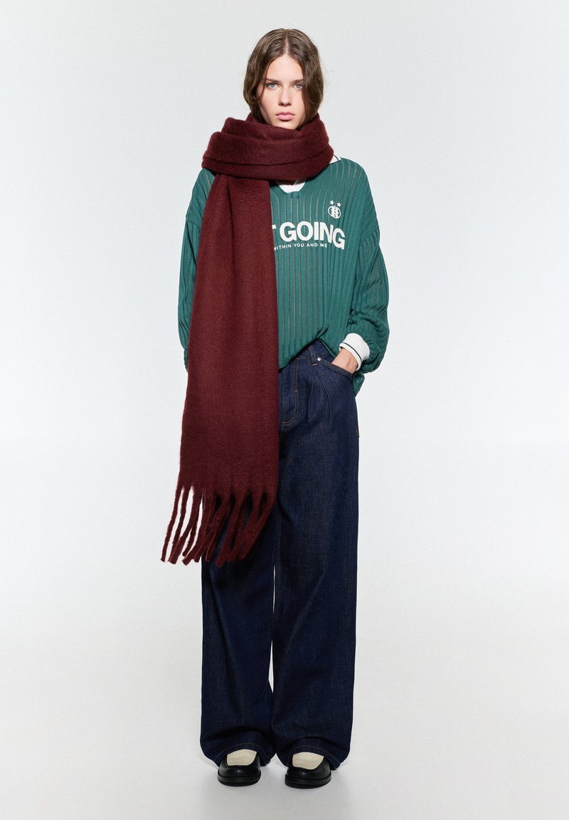 Vinskapad scarf i mjukt tyg med fransade ändar, kombinerad med en grön ribbad tröja och lösa mörkblå jeans. Svarta skor kompletterar outfiten.