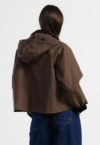 Giacca impermeabile marrone con cappuccio e fodera a quadri; presenta una vestibilità comoda e polsini a costine; abbinata a pantaloni di jeans blu.