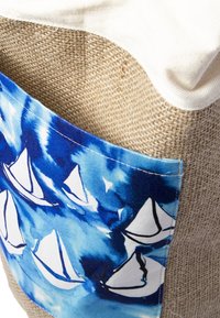 Nahaufnahme eines gewebten beigen Stoffs mit einer blauen Tasche, die weiße Segelbootmuster zeigt, und einem weichen, cremefarbenen Stoffrand.