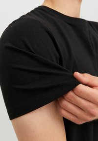 Fekete pamut póló rövid ujjakkal, kerek nyakkivágással és puha textúrával. Kép a kézről, amely a szál végén fogja a pólót.