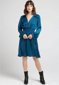 Robe bleu-vert avec un décolleté en V et une taille smockée, dotée de longues manches avec des poignets à volants. Portée avec des bottines noires. Texture lisse.