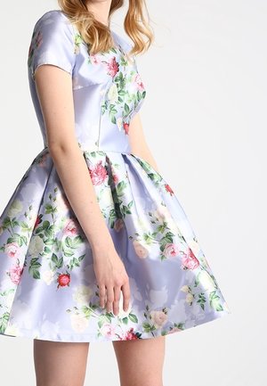 Kobieta w lawendowej sukience z krótkim rękawem do kolan, z różowo-białym kwiatowym wzorem, dopasowanym gorsetem i rozkloszowaną spódnicą na białym tle.