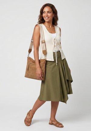 Femme portant un gilet sans manches crème noué devant, une jupe asymétrique vert olive, un sac bandoulière beige et des sandales beiges, debout et souriante.