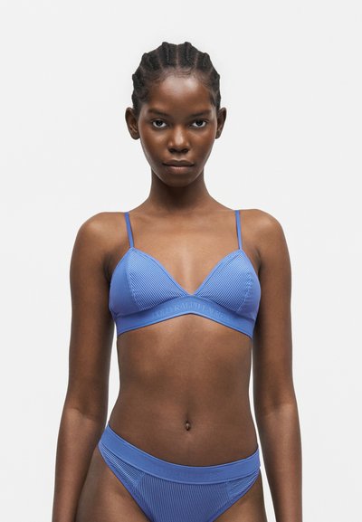 Γυναίκα με πλεγμένα μαλλιά που φοράει ταιριαστό μπλε ριμπ μπραλέτα και μπικίνι με ψηλή κοπή, κοιτάζοντας μπροστά σε ένα απλό φόντο.