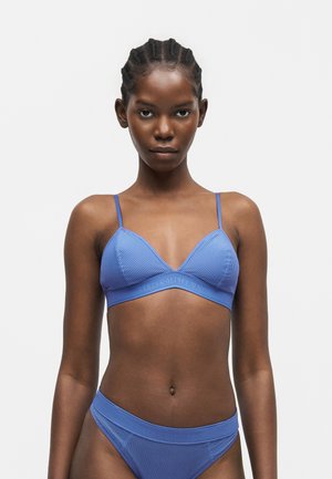 Kvinde med flettet hår iført en matchende blå ribbet bralette og bikinitrusser med høj benskæring, vendt fremad mod en ensfarvet baggrund.