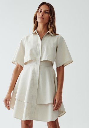 Licht beige jurk met korte mouwen, een rits aan de voorkant en een gelaagde, uitlopende rok. Voorzien van een omgeslagen kraag en een soepele stoftextuur.