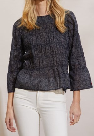 Kvinde iført en mørkeblå bluse med paisley-mønster og trekvartlange puffede ærmer samt hvide tætsiddende bukser mod en ensfarvet baggrund.