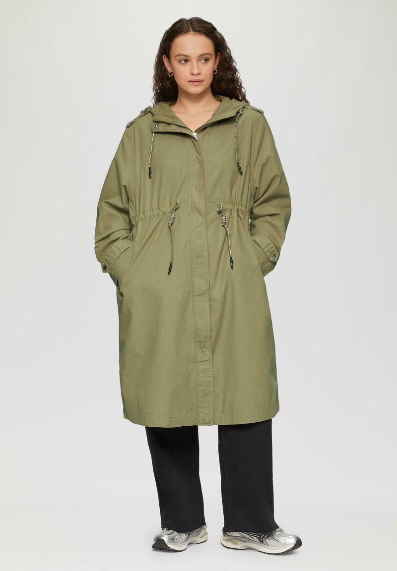 Vrouw met krullend haar, gekleed in een olijfgroene gewatteerde parka met capuchon, zwarte wijde broek en zilveren sneakers, staande met haar handen in de zakken.