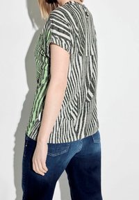 Gestreiftes Kurzarm-Oberteil in Grün und Grau, mit einem runden Ausschnitt und einem Knopfdetail auf der Rückseite. Kombiniert mit dunkelblauen Jeans.