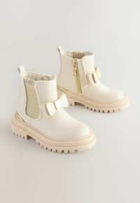 Weiße Knöchelstiefel mit glänzender Oberfläche, ausgestattet mit einer strukturierten Seitenpartie, einem Schleifenakzent und einem metallischen Reißverschluss an der Außenseite. Robuste Gummisohle.