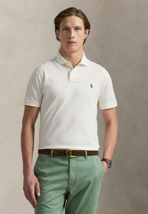 Young man wearing an off-white polo shirt with a small logo, green pants, brown belt, and wristwatch, standing against a plain background.