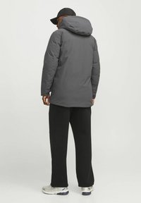 Grå polstret jakke med hætte, iført over sorte bukser. Har en glat tekstur og en afslappet pasform, parret med grå og sølv sneakers.