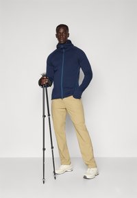 Marineblå zip-up hoodie med kontrasterende blå syninger, beige bukser og hvide sneakers. Holder sorte trekkingstænger mod en ensfarvet baggrund.