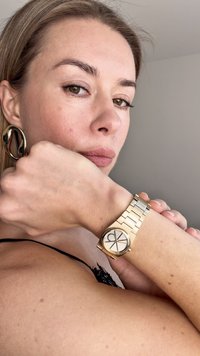 Aukso rankinis laikrodis su apvaliu ciferblatu ir juodais ženklais, turintis metalinį dirželį. Didelė auksinė auskarų kilpa kairėje ausyje, odos tekstūra glotni.