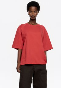 Camiseta roja de gran tamaño con mangas ragl�án cortas, fabricada en tejido suave. Presenta un escote redondo y un ajuste relajado, combinada con pantalones oscuros.