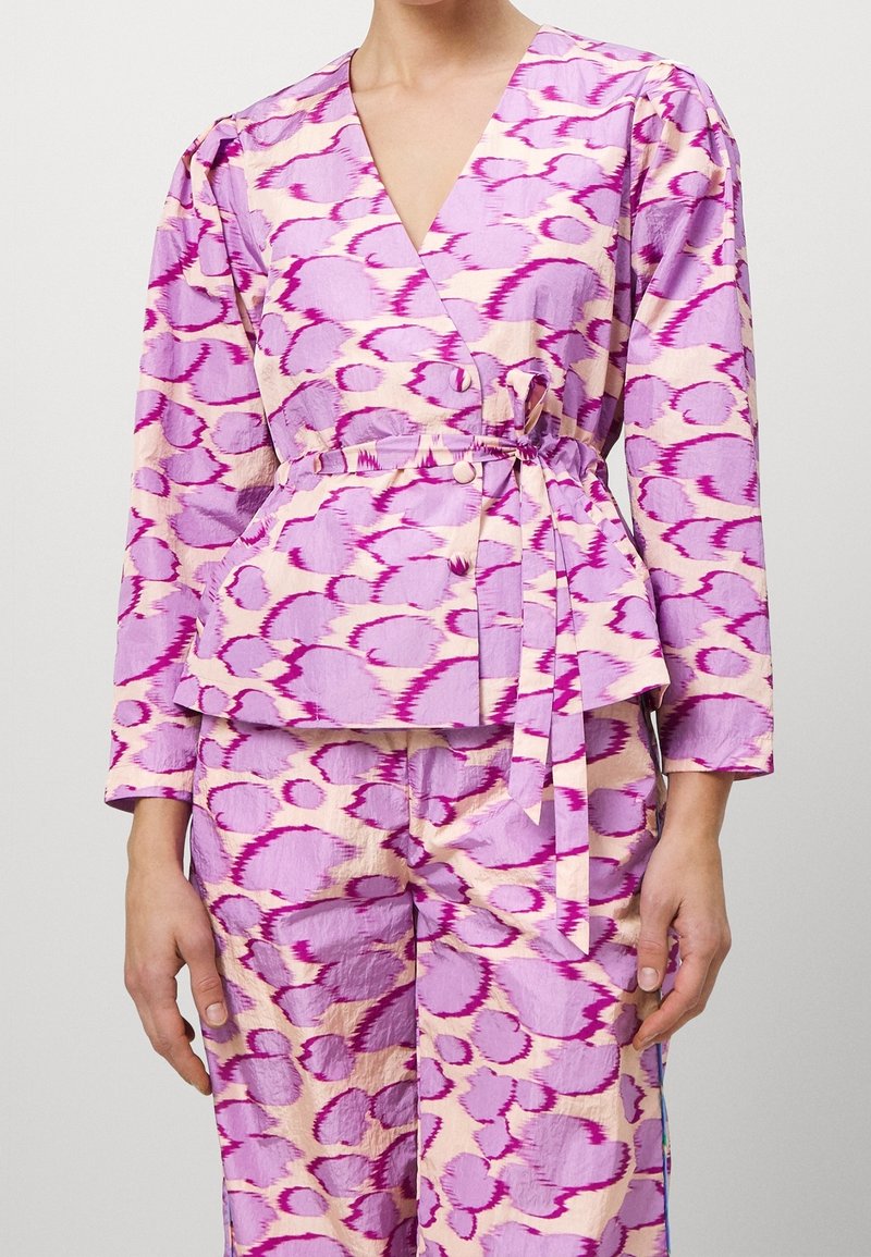 Femme portant un ensemble composé d'un haut cache-cœur à manches longues et d'un pantalon avec un motif circulaire abstrait violet et rose, avec une ceinture en tissu nouée à la taille.
