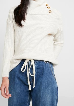 Kvinde iført en cremefarvet strikket sweater med knaplukket rullekrave og blå denim bukser med bindebånd foran, stående foran en ensfarvet baggrund.