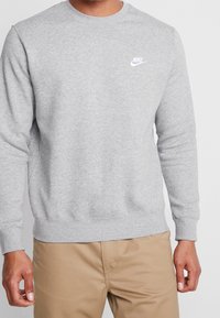 Sudadera gris hecha de una mezcla de algodón, con cuello redondo, mangas largas y un pequeño logotipo de Nike en blanco en el pecho. Tiene puños y dobladillo de canalé.