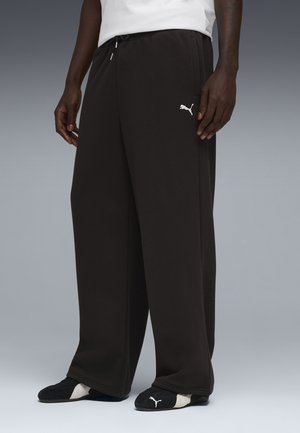 Sorte bredbenede Puma joggingbukser med snøre i taljen, båret af en stående person iført sorte og hvide Puma sneakers.