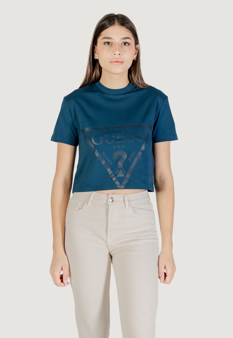 T-shirt cropped blu navy con un logo triangolare lucido nero che presenta la scritta "GUESS" e un testo grafico. Abbinato a jeans beige a vita alta.