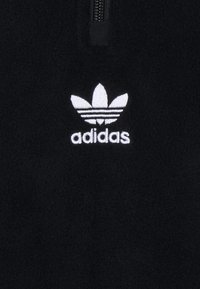 Svart fleece-tröja med kvartszipp och vit broderad Adidas trefoil-logga på vänster bröst. Mjuk textur och minimalistisk design.